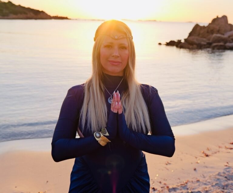Woman in a dark wetsuit on the beach at sunset with hands pressed together in prayer or meditation.